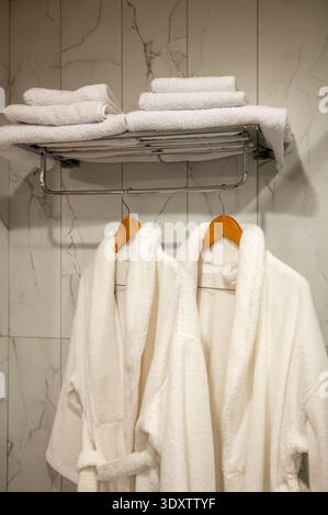 A clean and luxurious hotel bathroom scene featuring plush white terry cloth robes and neatly folded towels on a metal rack against a modern white mar Stock Photo