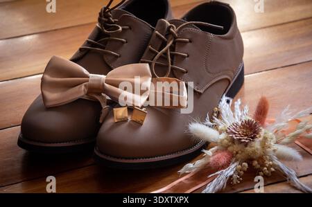 Close-up of groom accessories including brown leather dress shoes, bow tie and cufflinks arranged on a wooden floor with dried floral boutonniere. Stock Photo