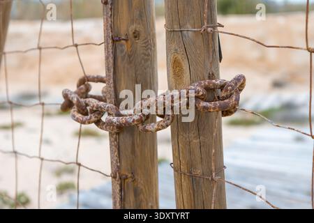 A weathered, rusted chain is tightly wrapped around two wooden posts, forming a sturdy, rustic fence against the backdrop of a sandy landscape. Stock Photo