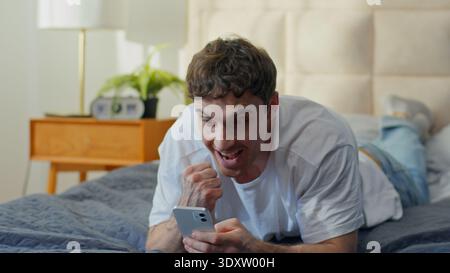 Happy Caucasian man lying on bed at home holding smartphone rejoicing victory win luck smiling joyful male guy reading good news message success Stock Photo