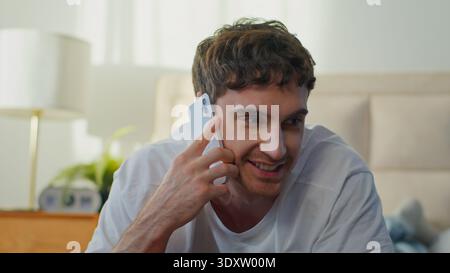 Happy laughing young Caucasian man talking on smartphone in bedroom European cheerful glad male guy discussing mobilr phone call indoor home friendly Stock Photo
