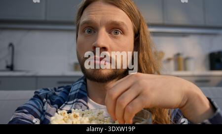 Concentrated Caucasian bearded man guy watching TV at home couch eating popcorn focused relaxed male enjoy series movie sports championship football Stock Photo