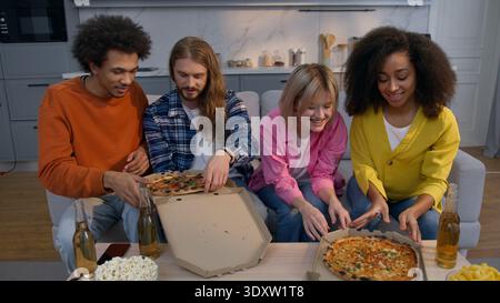 Food delivery service multiracial friends men women enjoying pizza at home celebrate party African Caucasian buddiess laughing talking eating on couch Stock Photo