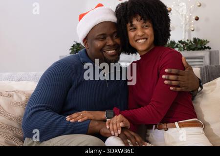 African American couple sitting on sofa at home, smiling, man wearing Santa hat holding hands Stock Photo