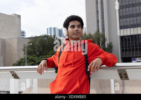 Teenage male leaning on rail at elevated walkway, wearing red hoodie, white headset, black backpack Stock Photo