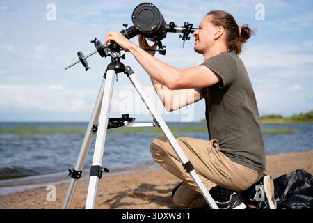 A man is looking into the eyepiece of a telescope while setting it up on a sunny beach. He carefully adjusts the view to observe the sun, demonstrating his interest in astronomy and solar phenomena. Stock Photo