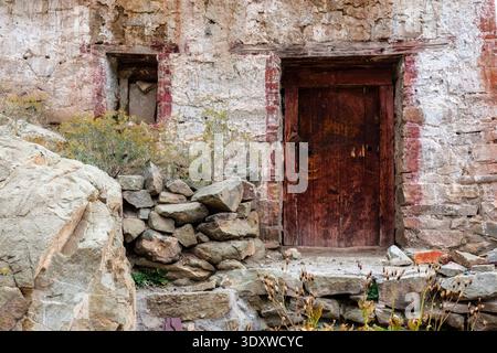 A weathered red wooden door and a small square window are set into the textured, whitewashed stone walls of Hemis Gompa in Hemis. Stock Photo