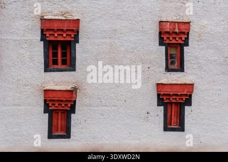 Four traditional small windows with red frames and black borders are arranged symmetrically on a white textured wall of Hemis Gompa. Stock Photo