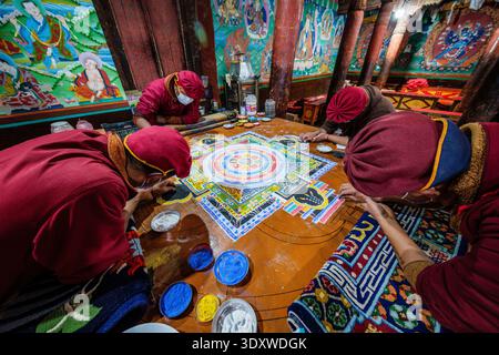 Several monks in traditional robes and face masks carefully assemble a complex sand mandala on a flat surface within the Hemis Gompa. Stock Photo