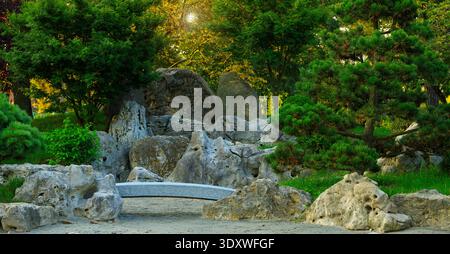A tranquil landscape featuring weathered rocks, a small arched bridge, and vibrant foliage illuminated by warm sunbeams filtering through the trees Stock Photo