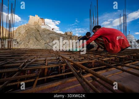 Two men tie steel reinforcement bars on a construction site in Leh, with the historic Leh Palace visible on a rocky hill in the distance. Stock Photo