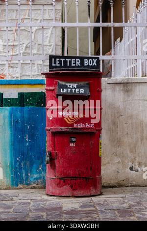 Red India Post pillar box standing on a paved street in Leh in front of a white metal fence and a wall with blue and green paint. Stock Photo