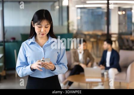 Focused East Asian businesswoman holding smartphone, standing in coworking office Stock Photo