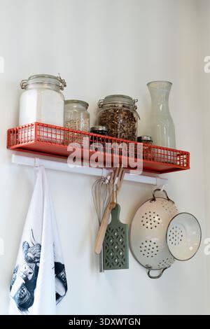Red metal kitchen shelf hanging on a white wall with jars of nuts and flour, a towel, strainer, and whisk vertical photo Stock Photo