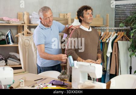 Elderly man tailor takes body measurements from adult man Stock Photo