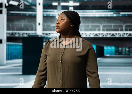 Woman walking through server hub rows lined with storage equipment rigs. African american IT worker inspecting high tech data center helping power internet services for business clients Stock Photo