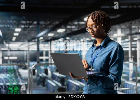 Data center black woman checking lines of code on laptop while she stands on industrial platform, overseeing AI tasks for automation. Server room IT expert visualizing deep learning process. Stock Photo