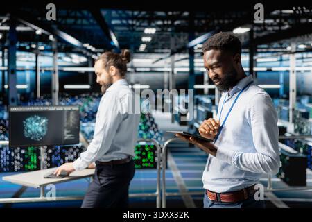 Software engineer next to colleagues reviewing neural network code and LLM visualization dashboards. Data center IT expert monitoring system performance with artificial intelligence tools. Stock Photo