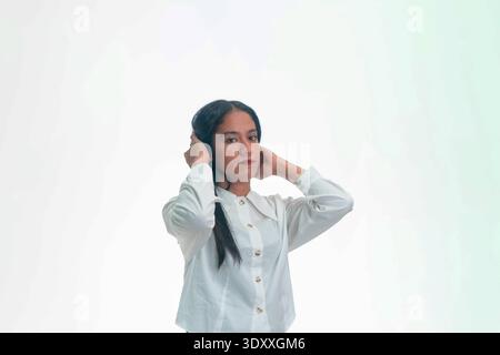 A young Indonesian woman in a white button-up shirt holding her hair with both hands in a preparation gesture, isolated on white studio background. Stock Photo