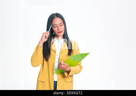 Thoughtful Indonesian woman in yellow blazer holding clipboard and touching head with pen, posing in a thinking gesture on white studio background. Stock Photo