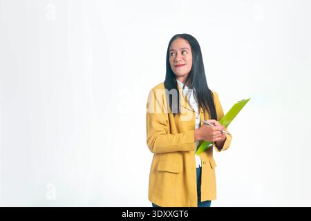 Happy Indonesian woman in yellow blazer holding a green clipboard and pen while looking up at copy space, isolated on white studio background. Stock Photo