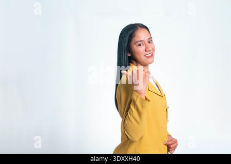 Cheerful Indonesian woman in yellow blazer looking at camera while pointing her thumb back at copy space, isolated on white studio background. Stock Photo