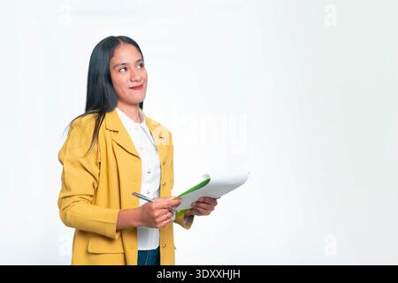 Professional Indonesian woman in yellow blazer holding a green clipboard and pen while looking sideways at copy space on white studio background. Stock Photo