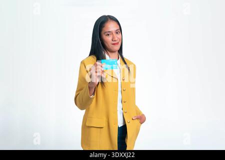 Portrait of a smiling Indonesian woman in a yellow jacket showing a blue credit card, isolated on white background for financial concepts. Stock Photo