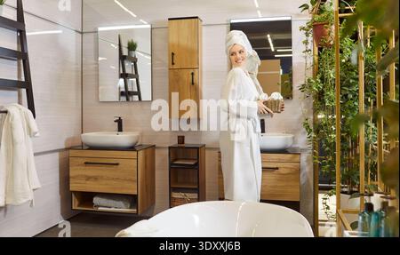 Woman happy standing in modern, spacious bathroom, smiling wearing white robe and towel wrapped Stock Photo