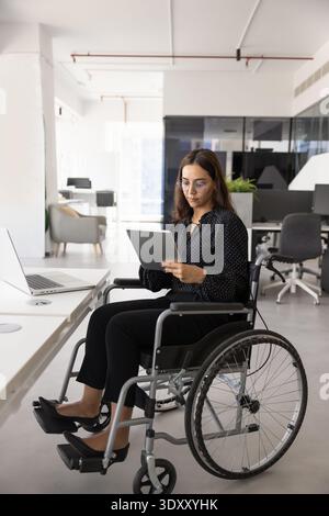 Serious young businesswoman in wheelchair working on online product Stock Photo