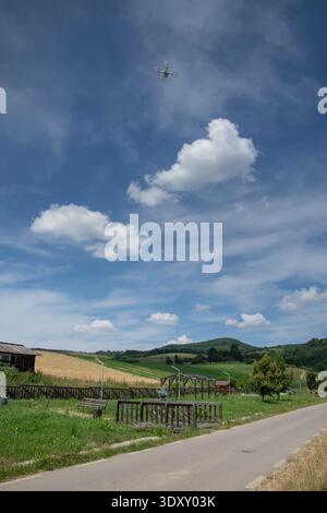 a drone flying over a rural countryside landscape with a road, green fields, and hills under a blue sky with clouds.  Drone: A small unmanned aerial v Stock Photo
