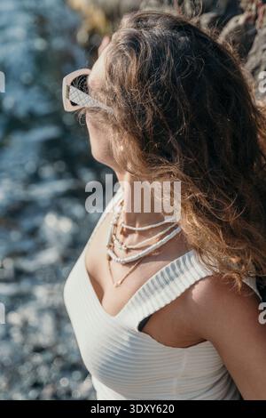 Woman sunglasses beach young lady in white summer top wearing necklaces enjoys sea view at rocky coast Stock Photo