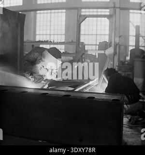 Authentic black and white archival image showcasing the intense labor of welders inside the bustling Stroymash plant in Sloviansk during the nineteen sixties. A skilled worker in a protective mask leans over a metal structure, generating a brilliant flash of light and smoke from the welding arc, while a colleague assists nearby. This dramatic industrial scene highlights Soviet manufacturing power, raw manual labor, and the dedicated working class within the historical context of a peaceful Donbass before the war. Stock Photo