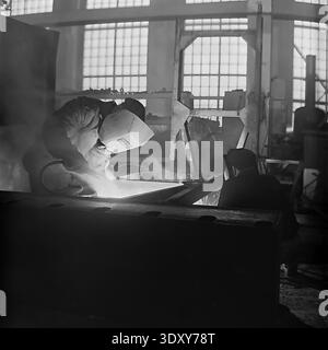 Authentic black and white archival image showcasing the intense labor of welders inside the bustling Stroymash plant in Sloviansk during the nineteen sixties. A skilled worker in a protective mask leans over a metal structure, generating a brilliant flash of light and smoke from the welding arc, while a colleague assists nearby. This dramatic industrial scene highlights Soviet manufacturing power, raw manual labor, and the dedicated working class within the historical context of a peaceful Donbass before the war. Stock Photo