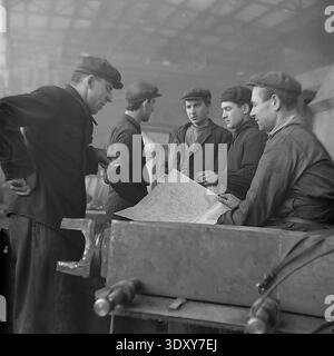 Impressive historical monochrome snapshot showing a group of five male factory workers actively discussing a technical blueprint at the Stroymash plant in Sloviansk in the 1960s. The skilled men, wearing traditional flat caps and heavy workwear, lean over a large paper plan resting on an industrial machine component. This engaging scene masterfully highlights Soviet engineering teamwork, collective intellectual labor, and the highly cooperative, constructive daily working environment of a peaceful Donbass before the war. Stock Photo