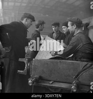Impressive historical monochrome snapshot showing a group of five male factory workers actively discussing a technical blueprint at the Stroymash plant in Sloviansk in the 1960s. The skilled men, wearing traditional flat caps and heavy workwear, lean over a large paper plan resting on an industrial machine component. This engaging scene masterfully highlights Soviet engineering teamwork, collective intellectual labor, and the highly cooperative, constructive daily working environment of a peaceful Donbass before the war. Stock Photo