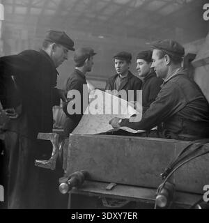 Impressive historical monochrome snapshot showing a group of five male factory workers actively discussing a technical blueprint at the Stroymash plant in Sloviansk in the 1960s. The skilled men, wearing traditional flat caps and heavy workwear, lean over a large paper plan resting on an industrial machine component. This engaging scene masterfully highlights Soviet engineering teamwork, collective intellectual labor, and the highly cooperative, constructive daily working environment of a peaceful Donbass before the war. Stock Photo