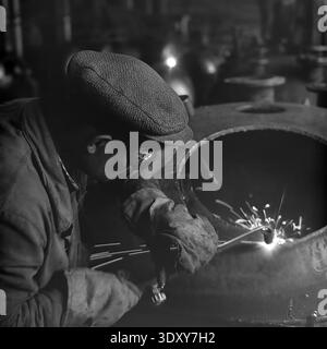 Striking archival monochrome image capturing a deeply focused male welder wearing protective goggles and heavy workwear engaged in intense labor inside the Stroymash plant in Sloviansk in the 1960s. The dedicated worker uses a bright cutting torch on a massive curved metal tank, generating brilliant sparks and blinding light against the dark factory workshop background. This dramatic and powerful composition masterfully showcases raw Soviet industrial effort, skilled manual craftsmanship, and the heartbeat of a peaceful Donbass before the war. Stock Photo