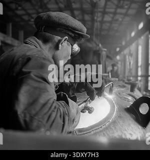 Striking archival monochrome image capturing a deeply focused male welder wearing protective goggles and heavy workwear engaged in intense labor inside the Stroymash plant in Sloviansk in the 1960s. The dedicated worker uses a bright cutting torch on a massive curved metal tank, generating brilliant sparks and blinding light against the dark factory workshop background. This dramatic and powerful composition masterfully showcases raw Soviet industrial effort, skilled manual craftsmanship, and the heartbeat of a peaceful Donbass before the war. Stock Photo