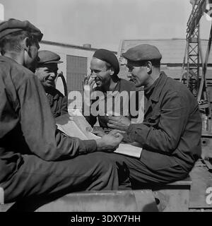 Fantastic archival monochrome photograph highlighting four cheerful male factory workers reviewing technical plans outdoors at the Stroymash plant in Sloviansk during the nineteen sixties. The happy comrades in flat caps sit together in the bright sunlight, smoking and actively discussing the paperwork with genuine smiles. This incredibly candid documentary snapshot brilliantly captures Soviet engineering teamwork, true workplace friendship, and the remarkably positive, constructive everyday working rhythm of a peaceful Donbass before the war. Stock Photo