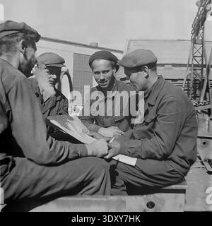 Fantastic archival monochrome photograph highlighting four cheerful male factory workers reviewing technical plans outdoors at the Stroymash plant in Sloviansk during the nineteen sixties. The happy comrades in flat caps sit together in the bright sunlight, smoking and actively discussing the paperwork with genuine smiles. This incredibly candid documentary snapshot brilliantly captures Soviet engineering teamwork, true workplace friendship, and the remarkably positive, constructive everyday working rhythm of a peaceful Donbass before the war. Stock Photo