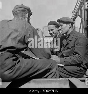Engaging historical monochrome picture capturing four male factory workers actively discussing technical blueprints outdoors at the Stroymash plant in Sloviansk during the 1960s. The cheerful comrades in flat caps sit together on a metal structure, smoking and sharing a friendly conversation in the bright daylight. This highly authentic documentary snapshot beautifully showcases Soviet engineering teamwork, genuine workplace friendship, and the positive, constructive social reality of a peaceful Donbass before the war. Stock Photo
