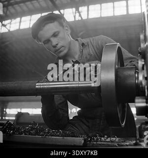 Striking archival black and white photograph highlighting a deeply concentrated young male machinist meticulously measuring a massive metal shaft with a caliper at the Stroymash plant in Sloviansk in the 1960s. Illuminated by beautiful dramatic light, the skilled worker in a flat cap carefully controls the precision turning process on a heavy lathe. This captivating industrial portrait perfectly embodies Soviet technical craftsmanship, individual dedication, and the productive daily rhythm of a peaceful Donbass before the war. Stock Photo