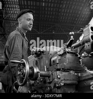 Striking archival black and white photograph highlighting a deeply concentrated young male machinist meticulously measuring a massive metal shaft with a caliper at the Stroymash plant in Sloviansk in the 1960s. Illuminated by beautiful dramatic light, the skilled worker in a flat cap carefully controls the precision turning process on a heavy lathe. This captivating industrial portrait perfectly embodies Soviet technical craftsmanship, individual dedication, and the productive daily rhythm of a peaceful Donbass before the war. Stock Photo