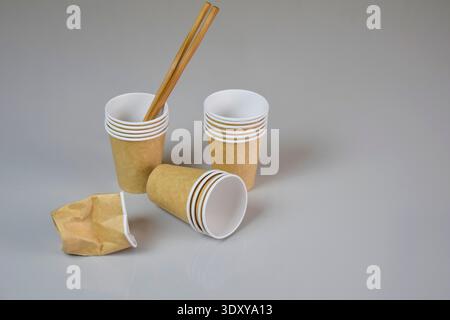 Brown disposable biodegradable cardboard cups for hot cold drinks and wooden sticks for stirring drinks on gray background. One paper cup is crumpled. Stock Photo
