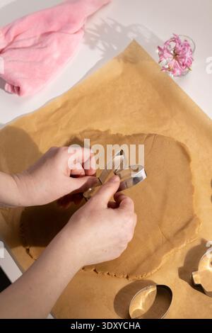 Hands are using a bunny shaped cookie cutter to cut out dough for gingerbread cookies. The setting is a kitchen with baking supplies and other cookie shapes nearby, preparing for Easter festivities. Stock Photo