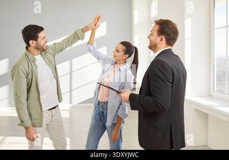 Young couple giving high five each other happy to buy a house standing with smiling man realtor. Stock Photo