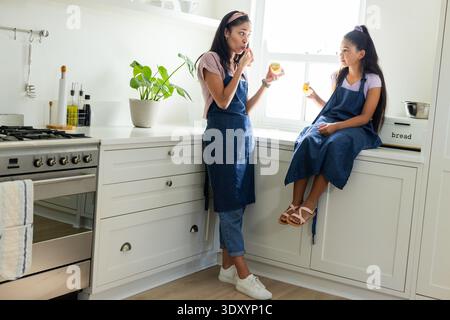 African American mom, youth daughter in denim aprons tasting from jars with spoon at home counter Stock Photo
