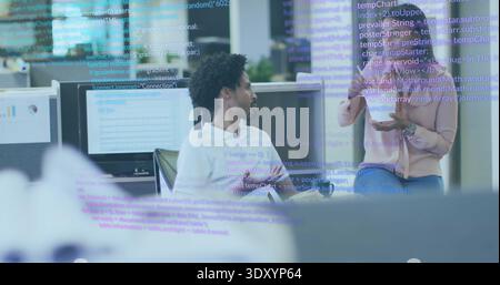 Talking two coworkers pointing at monitors in office, in business casual with tablet, code overlay Stock Photo