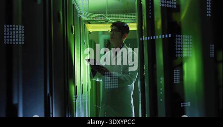 Inspecting Korean tech wearing white lab coat, holding tablet, checking server LEDs in data center Stock Photo
